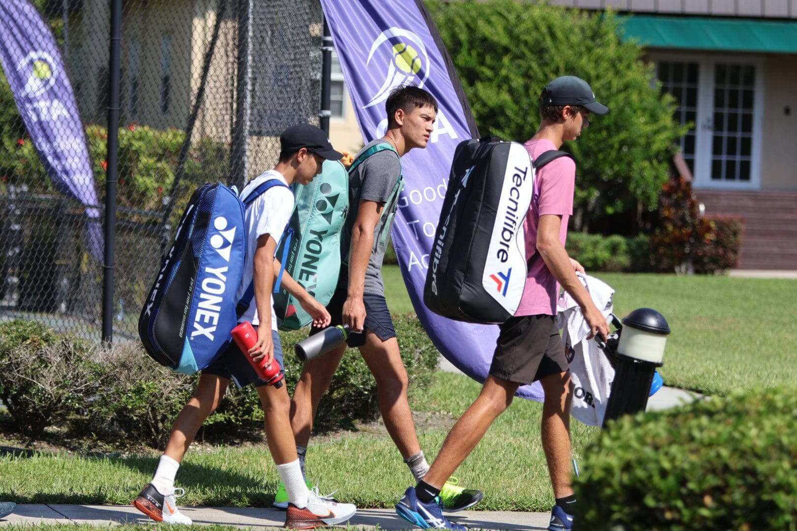 Three young men walking with tennis gear bags on a sunny day.