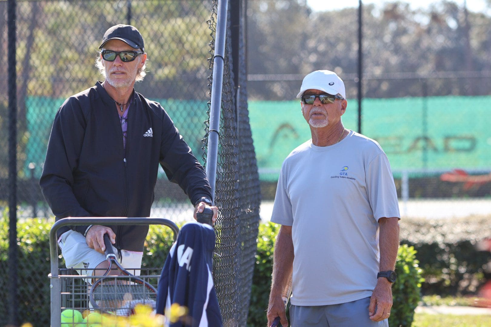 Two men standing near a tennis court, one holding a paddle.