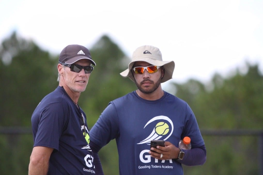Two men in sunglasses and hats stand outdoors, wearing matching navy shirts.