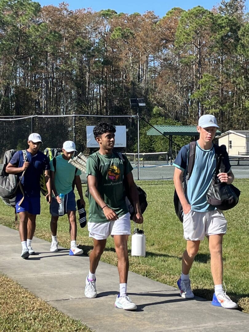 Four young men walking on a sunny day with tennis rackets and backpacks.