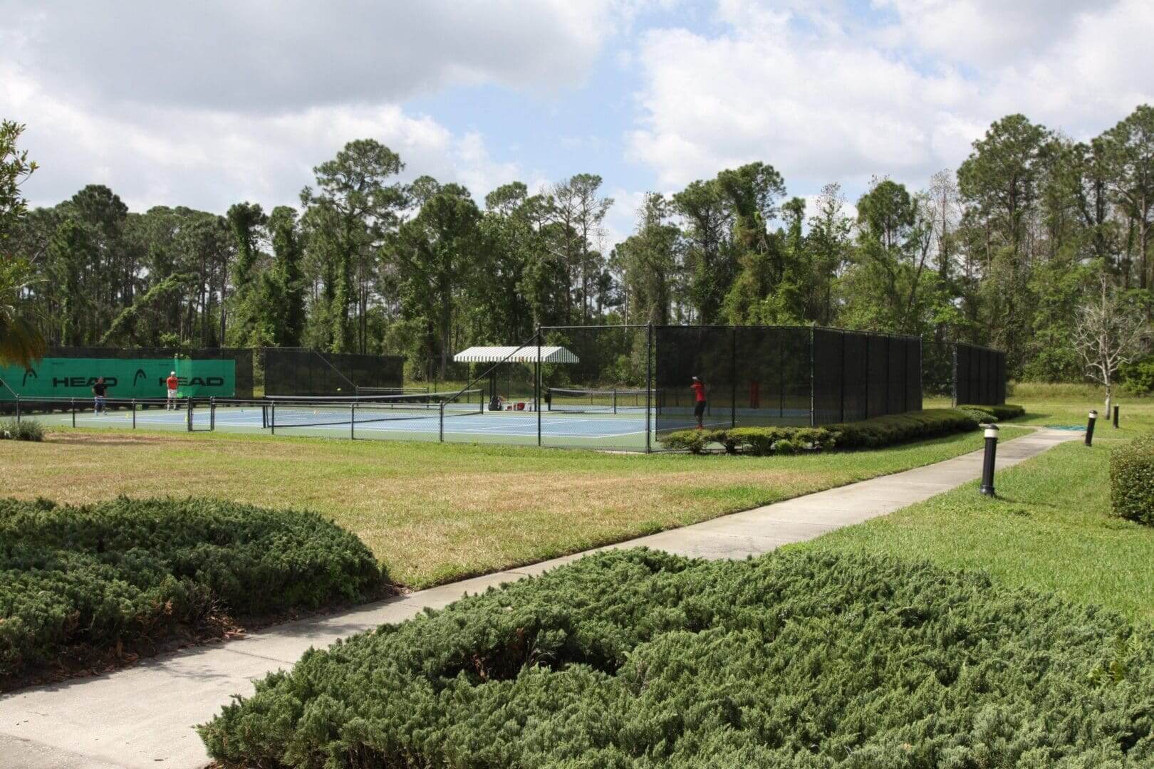 Outdoor tennis courts surrounded by trees and greenery under a cloudy sky.