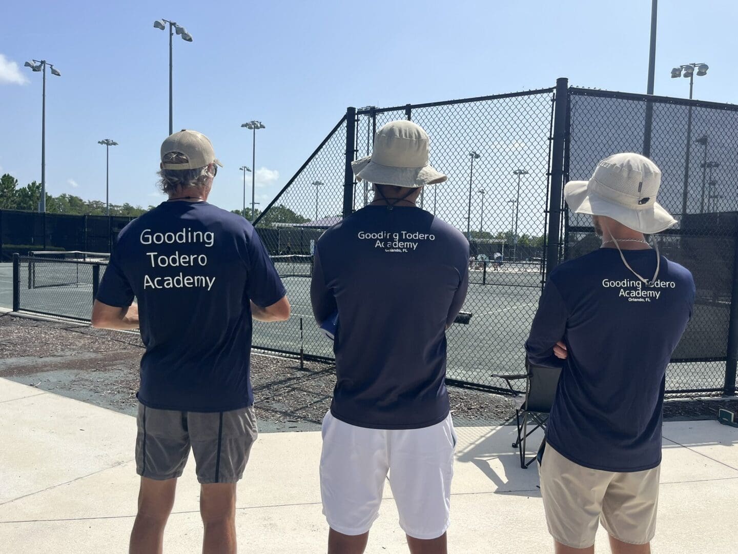 Three young men wearing 'Gentlemen's Academy' shirts stand by a tennis court.