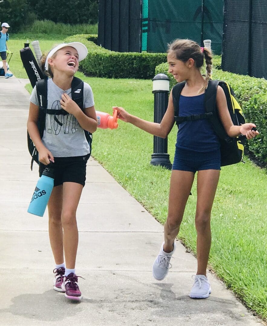 Two girls walking with tennis rackets, sharing a drink on a sunny day.