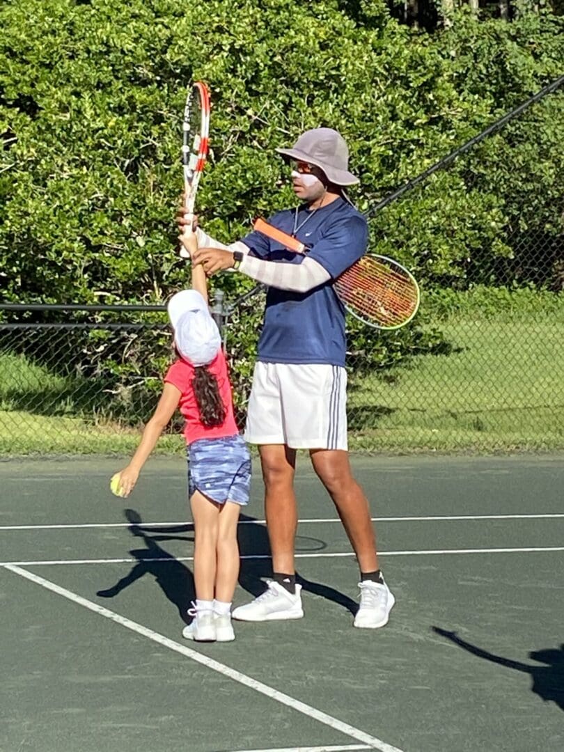 Adult teaching child tennis on outdoor court.